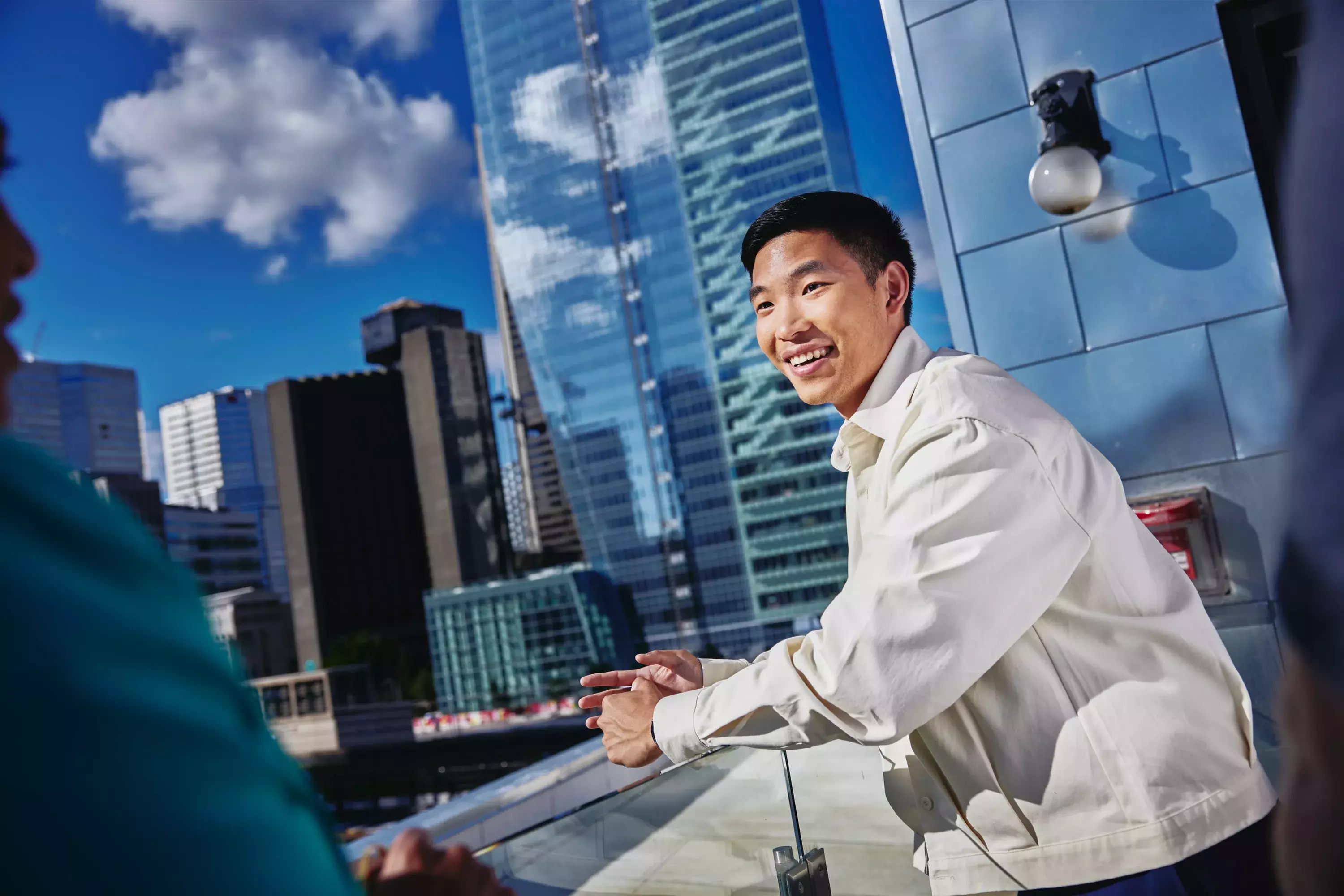 Male smiling, standing outside on a balcony talking to a person