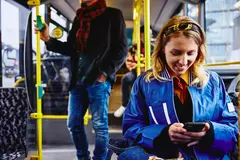 Smiling female sitting in a bus, looking at her phone. Other people in the background.
