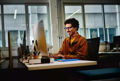 Male with blue glasses looking at a computer screen sitting at a desk